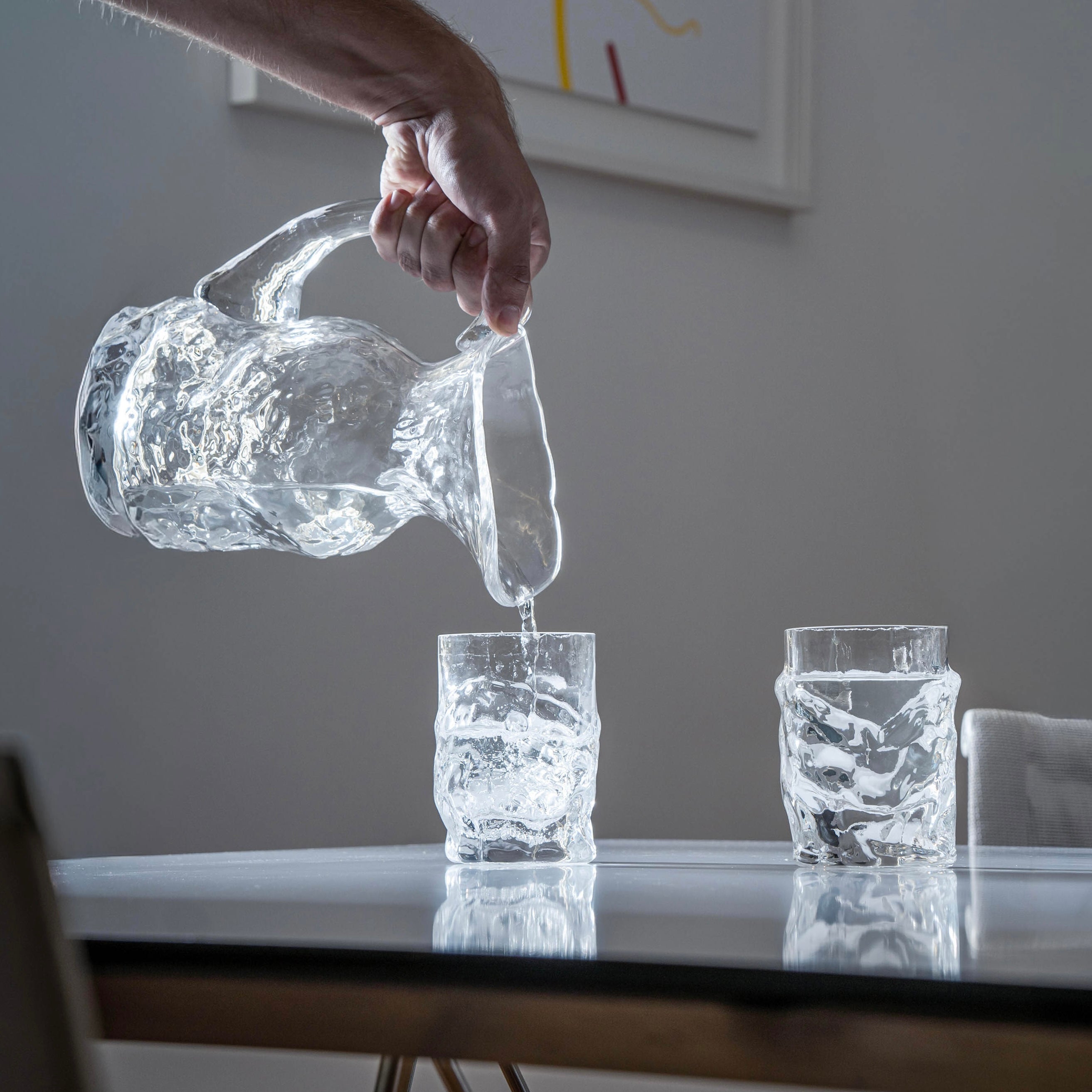Person pouring water from a clear glass pitcher into two glasses on a table with abstract art in the background.