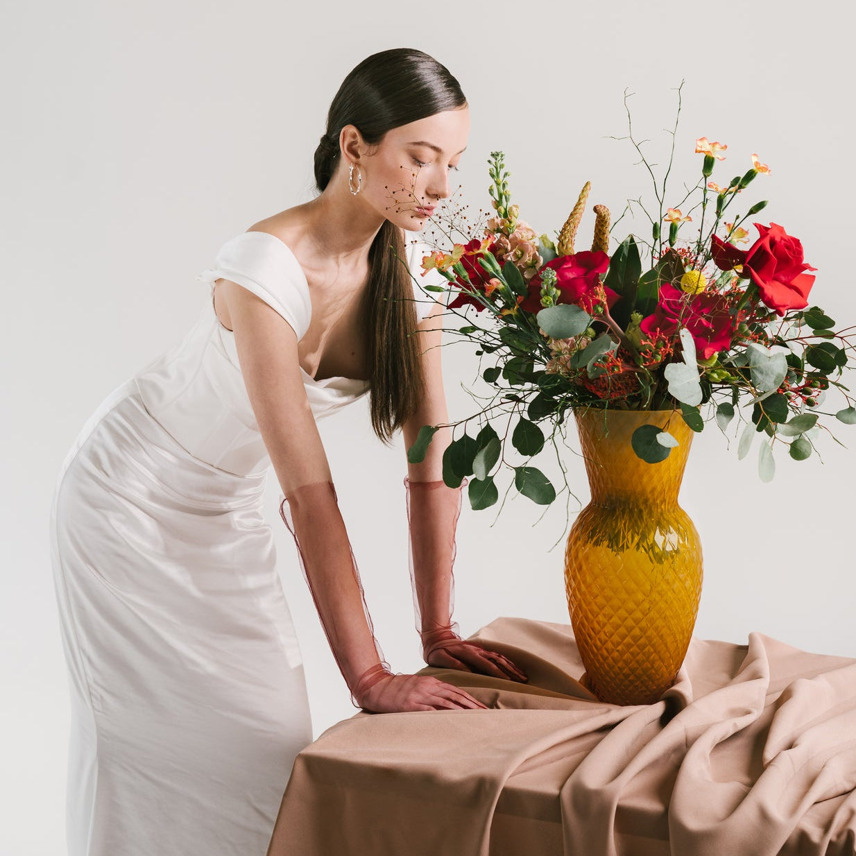 Woman in a white dress arranging flowers in a yellow vase on a table with a plain background