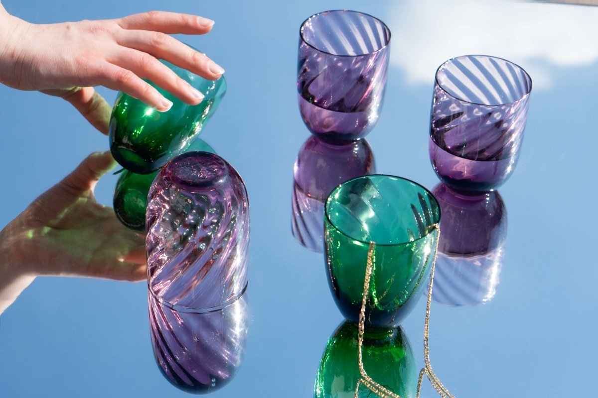 Violet Marika Tumblers on a mirror reflecting the sky, accompanied by gold jewellery and a moving hand
