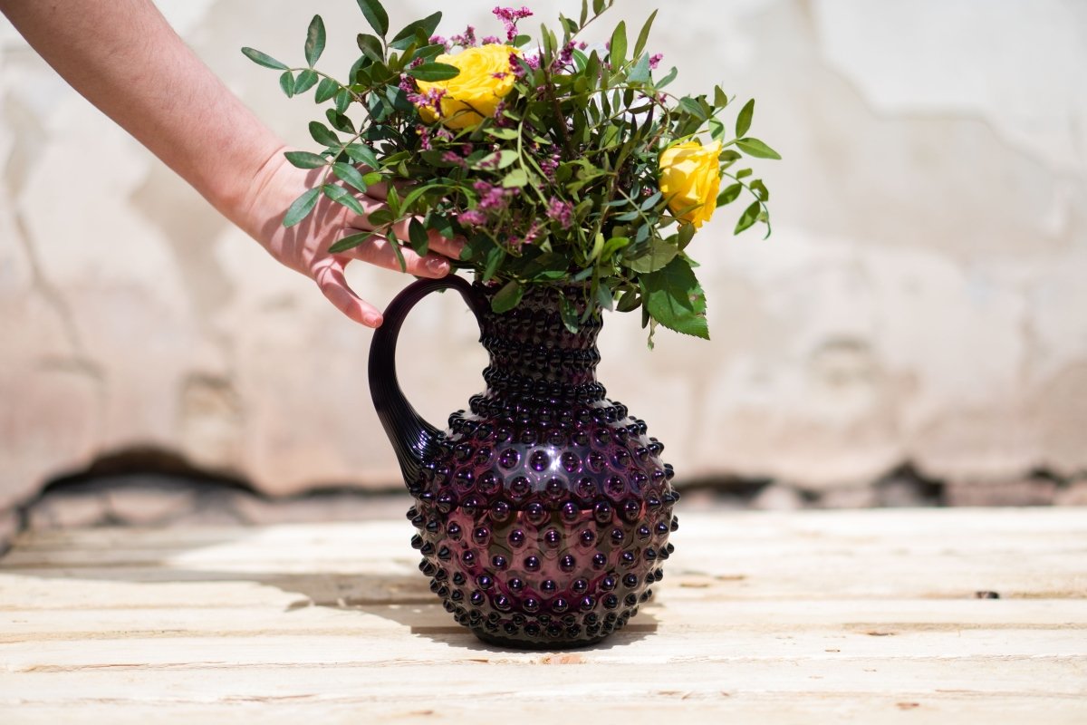 Violet Hobnail Jug on a wooden table with purple and yellow flowers and a broken wall in the background