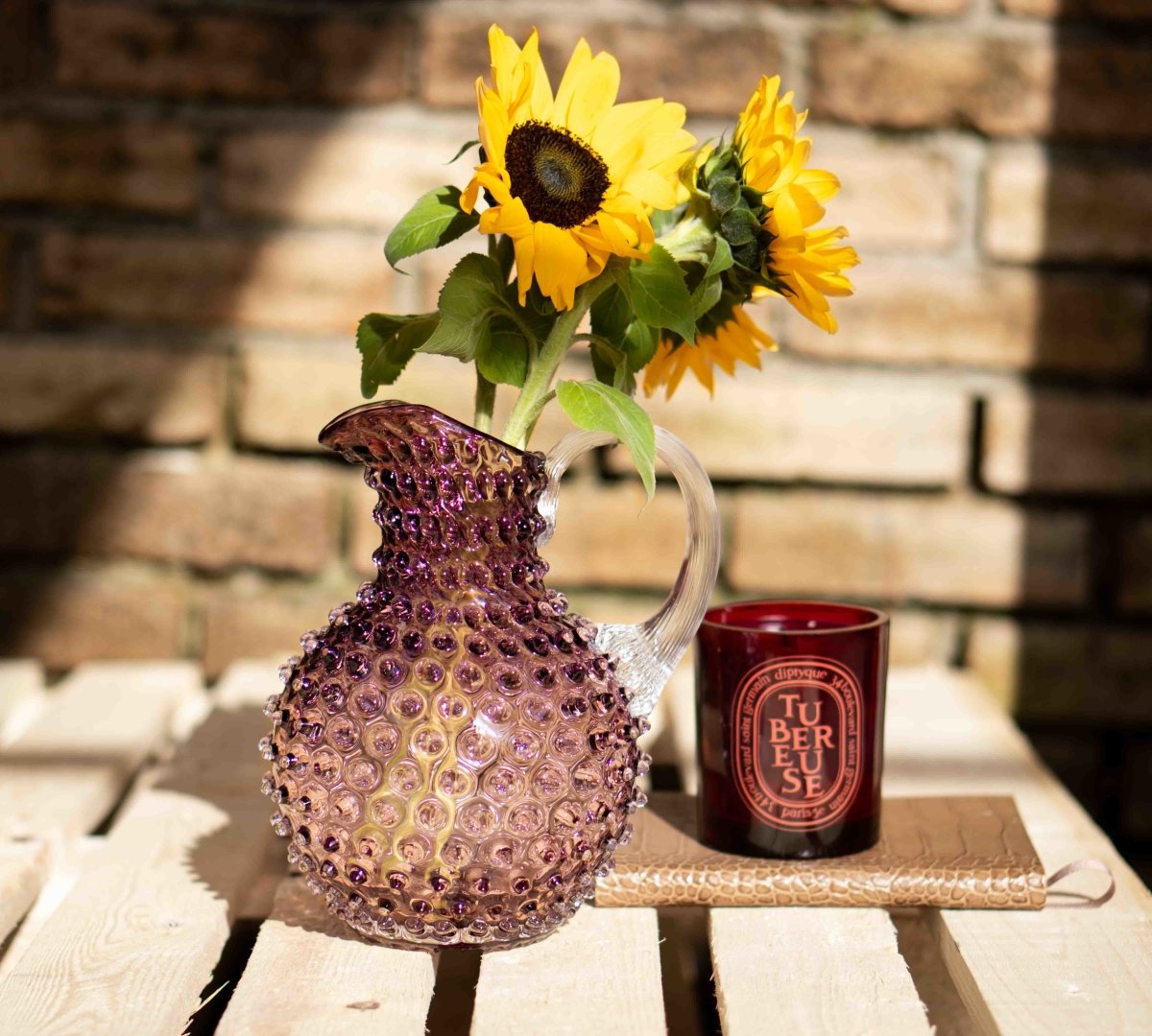 Underlay Violet Hobnail Jug on a wooden pallet with yellow flowers and a brick wall in the background
