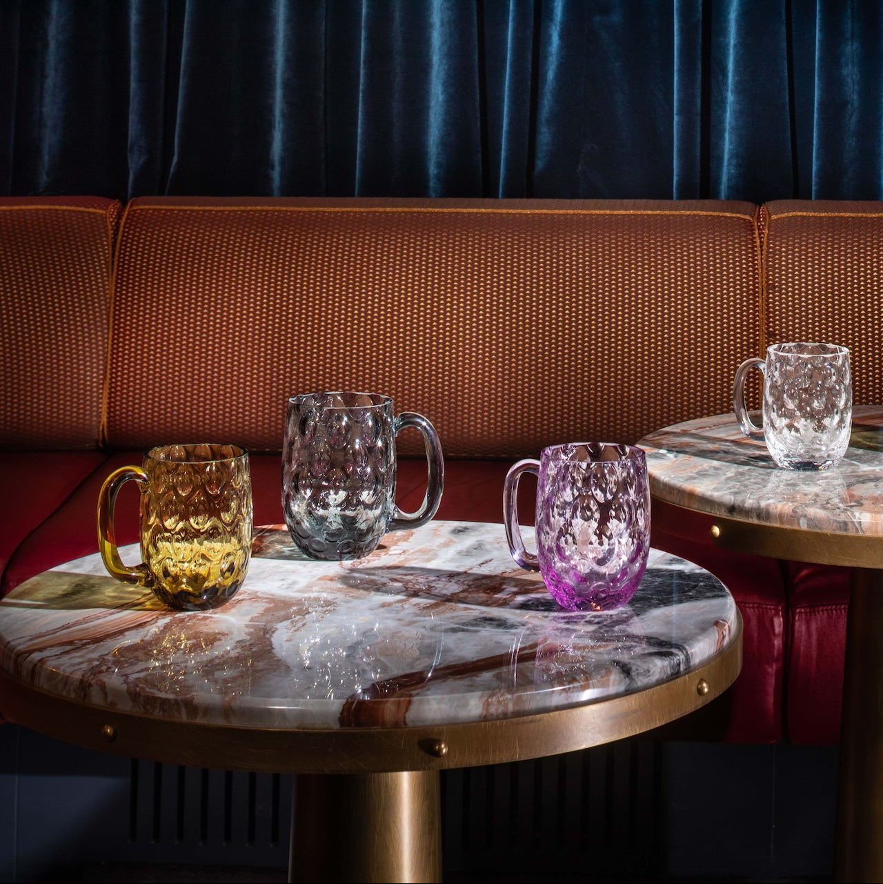 Two round tables with marble tops and gold bases, each holding colorful glass mugs, against a dark curtain background.