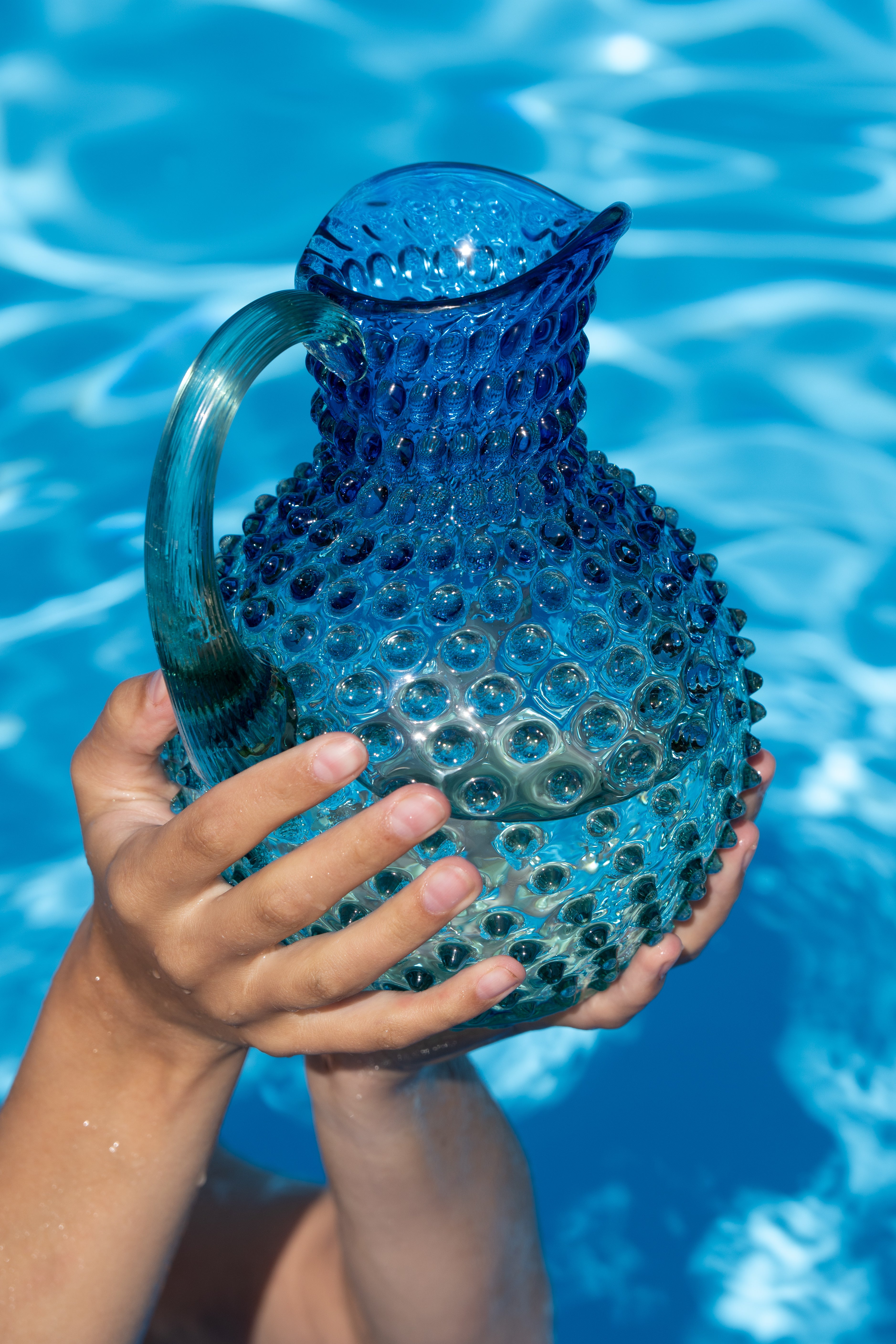 A translucent blue glass pitcher with a textured hobnail pattern, held by two hands in front of a bright blue swimming pool background.