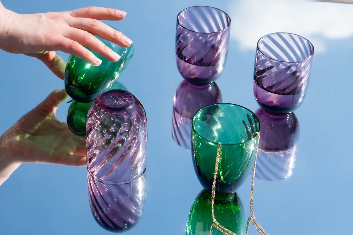 Violet Marika Tumblers on a mirror reflecting the sky, accompanied by gold jewellery and a moving hand 