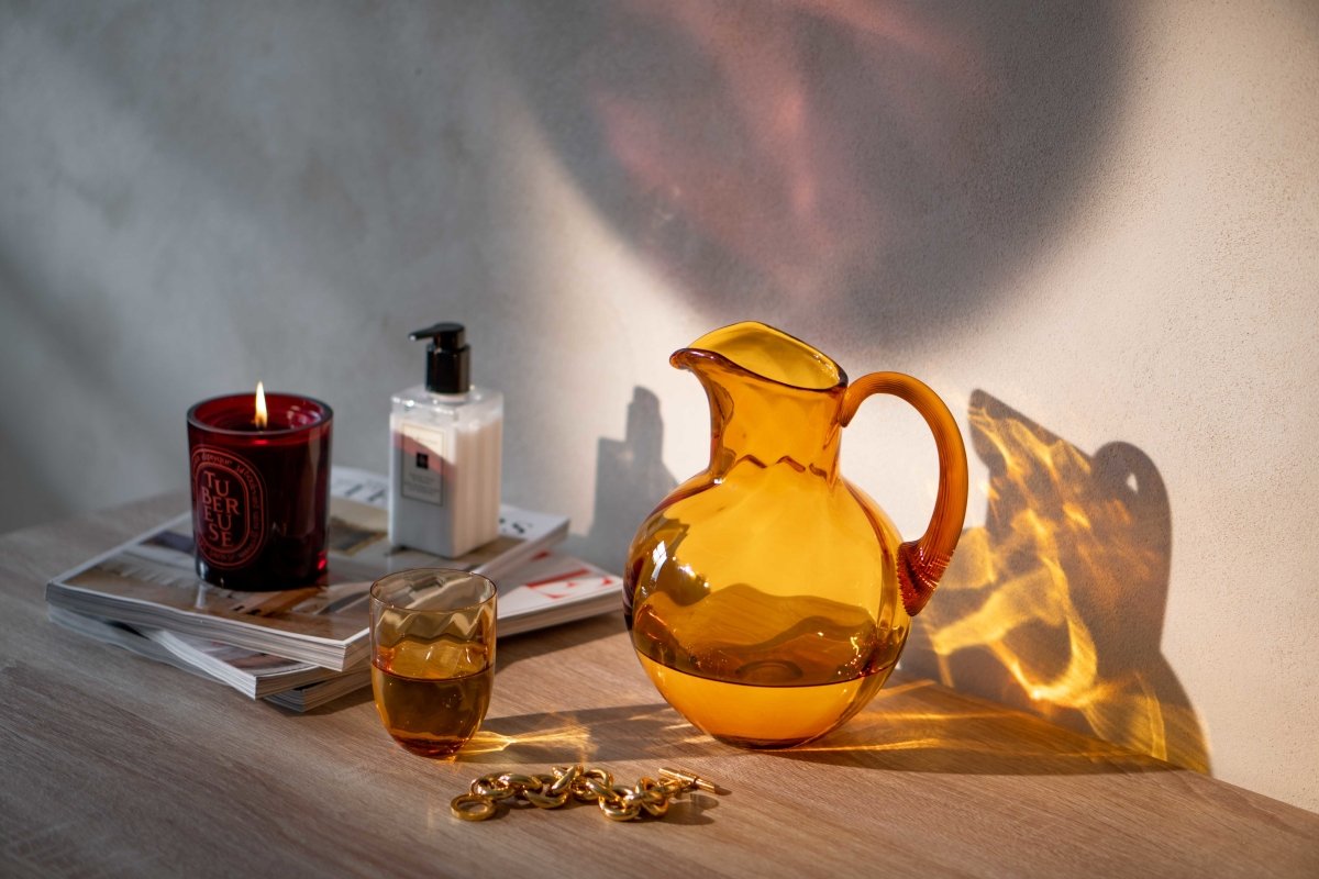 Amber Marika Tumblers on a wooden table surrounded by magazines, a beauty product, a candle, a necklace and a Marika Amber jug